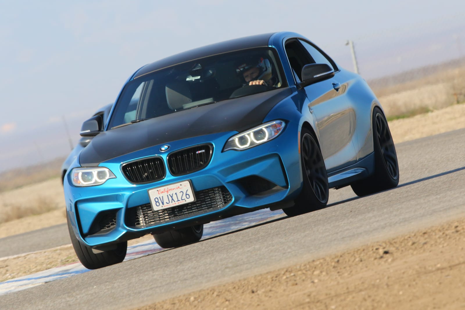 A blue BMW M2 with a black hood at speed on track at Buttonwillow Raceway Park.