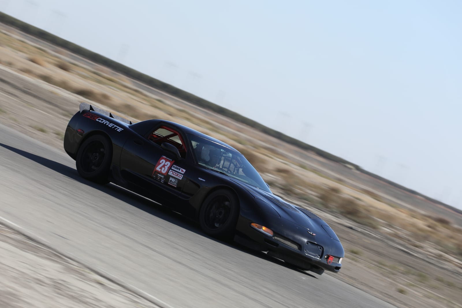 A black FastLane Spec Corvette panning at speed on track at Buttonwillow Raceway.