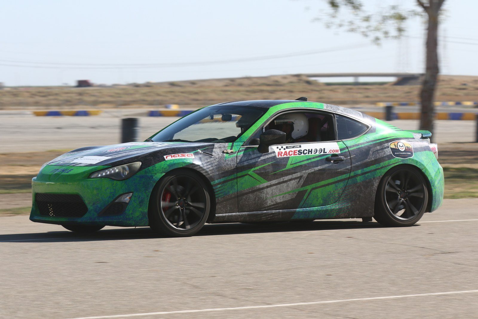 A green and black FastLane Toyota GR86 / FRS in RaceSchool.com livery cornering on track at Buttonwillow Raceway.