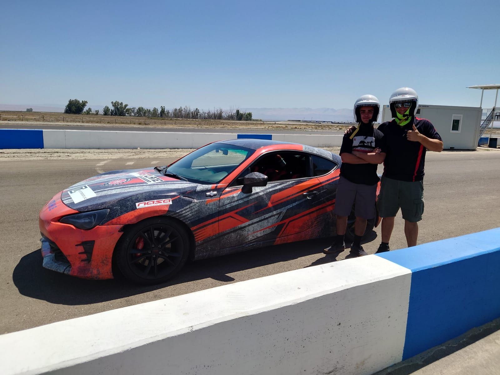 A FastLane instructor and a helmeted student beside a track-prepped car in the Buttonwillow pit.