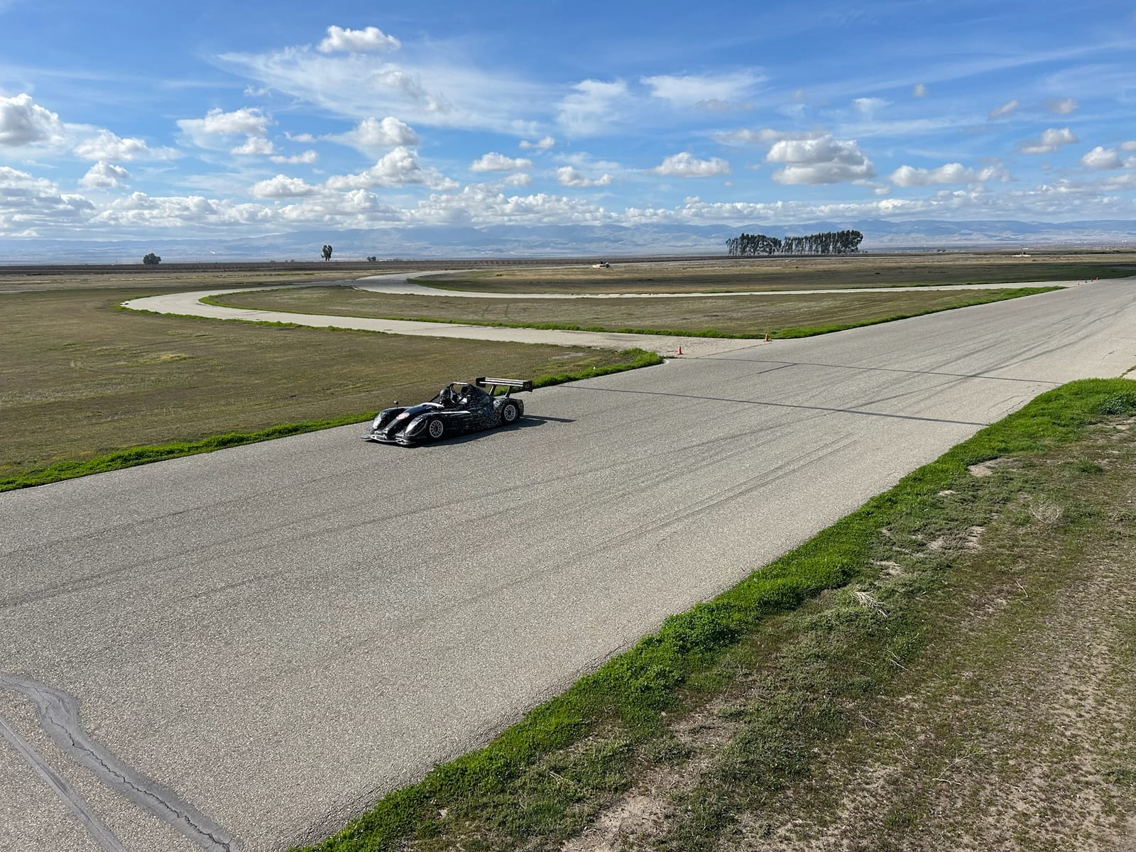 Elevated wide view of Buttonwillow Raceway with a race car mid-corner under a big cloud-filled sky.